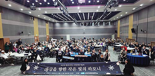 참석자들이 세상을 향한 작은 빛의 하모니라고 적혀 있는 현수막을 들고 단체 기념 촬영을 하고 있다